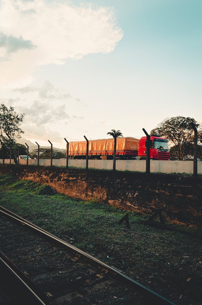 Red truck driving along a road beside railway tracks and trees in Resende, Brazil.