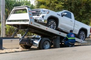 During the day, a flatbed truck driver loads a white GMC pickup truck onto the street.